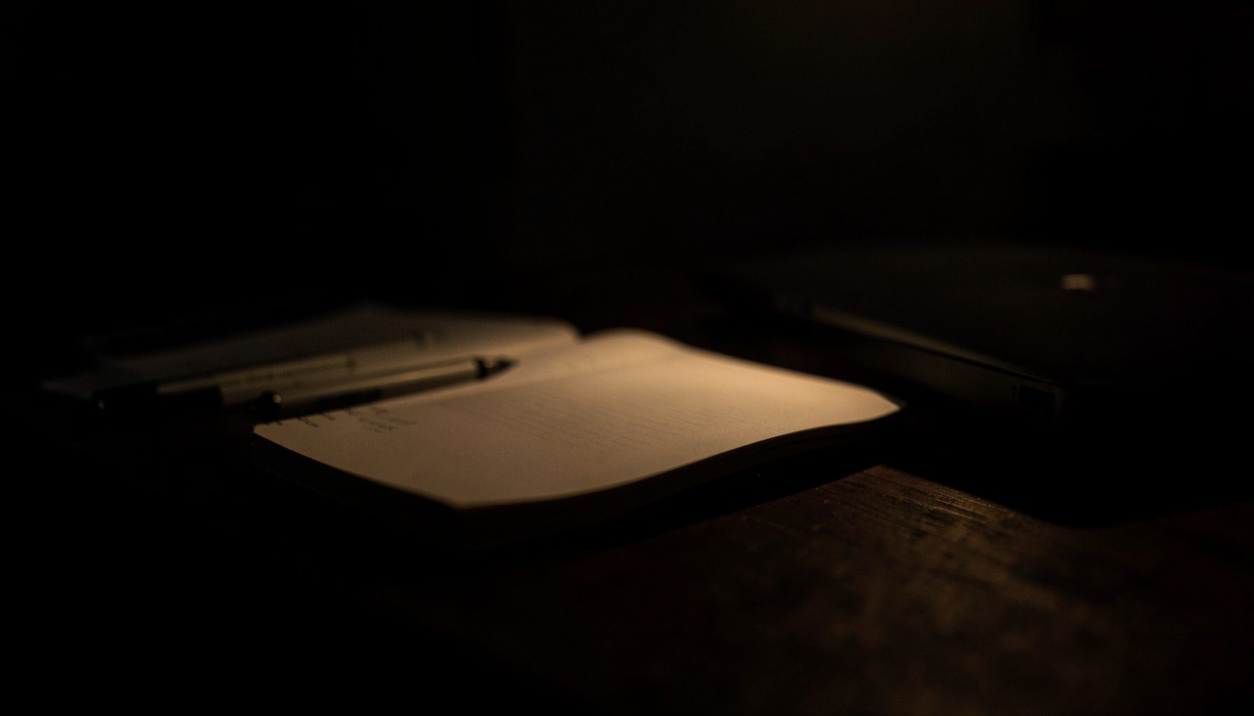 Mesa de trabalho escura com caderno aberto, caneta e um laptop parcialmente visível, iluminada por luz suave e quente