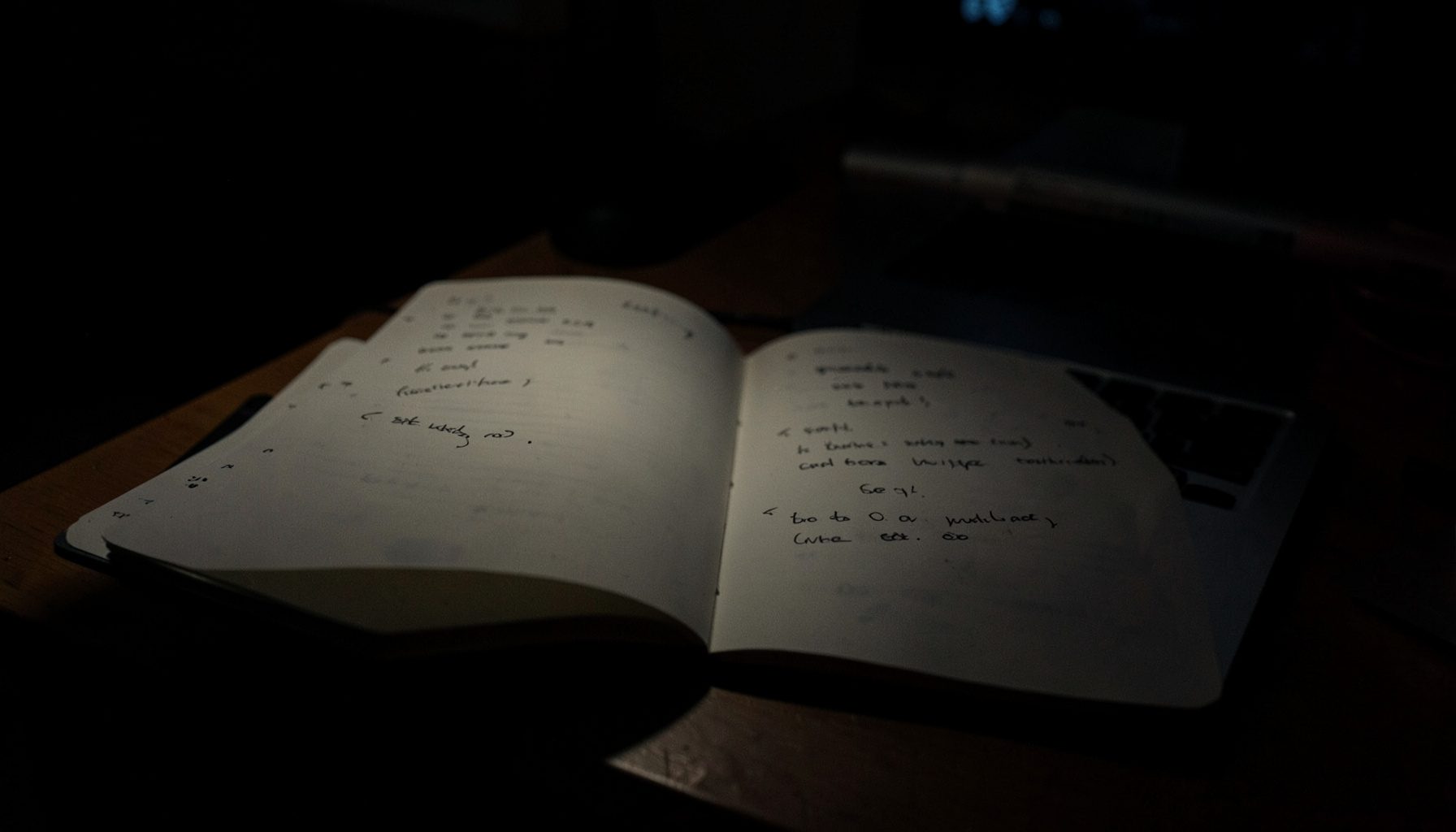 Caderno aberto com anotações ao lado de laptop, iluminação baixa e ambiente de trabalho noturno