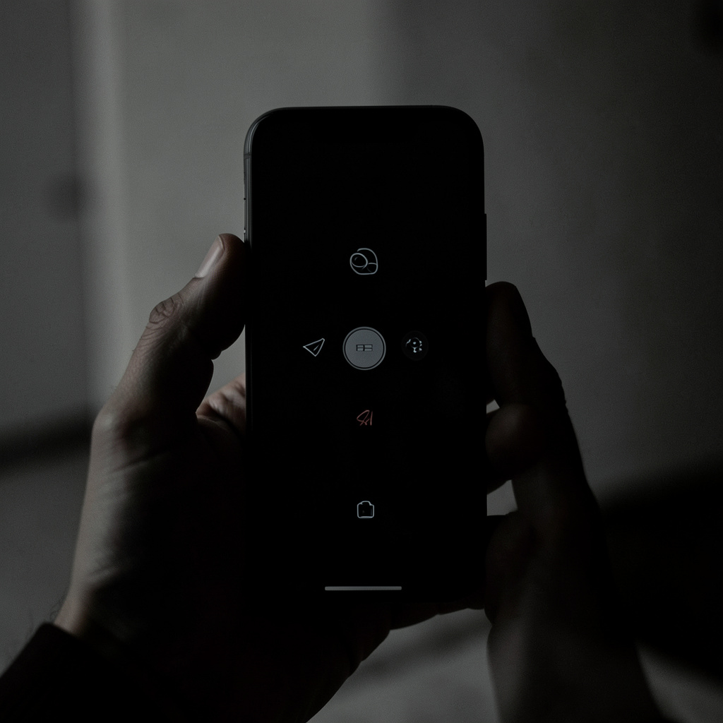 Mão segurando smartphone com tela escura exibindo ícones minimalistas, ambiente com pouca luz e tons de cinza e preto.